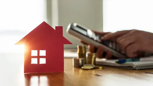 Baufinanzierung für Ihr Massivhaus in Lengerich Rotes Hausmodell aus Papier, daneben Münzstapel und Person mit Taschenrechner auf einem Holztisch.