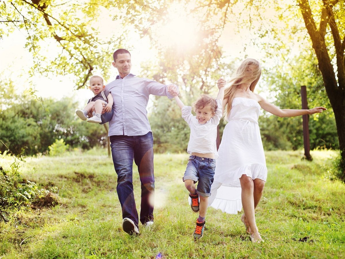 Familie mit zwei Kindern läuft glücklich durch eine grüne Parklandschaft im Sonnenlicht