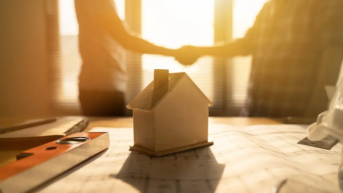 Hausbauplanung mit Beratung und Vertragsabschluss Hausmodell auf Bauplan mit Handschlag im Hintergrund als Symbol für Hausbauvertrag, Planung und Beratung