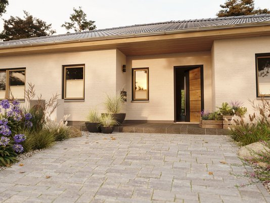Moderner ebenerdiger Bungalow von Thielker MassivHaus mit heller Fassade, Eingangstür, großer Pflasterfläche und bepflanztem Vorgarten.