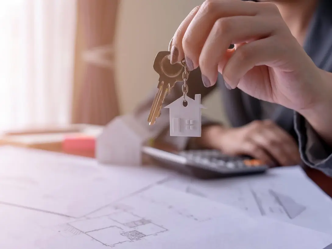 Von der Planung bis zur Schlüsselübergabe – Ihr Hausbau Schritt für Schritt. Hand hält Hausschlüssel über Bauplan und Taschenrechner – Symbolbild für Hausbau, Planung und Eigenheim.