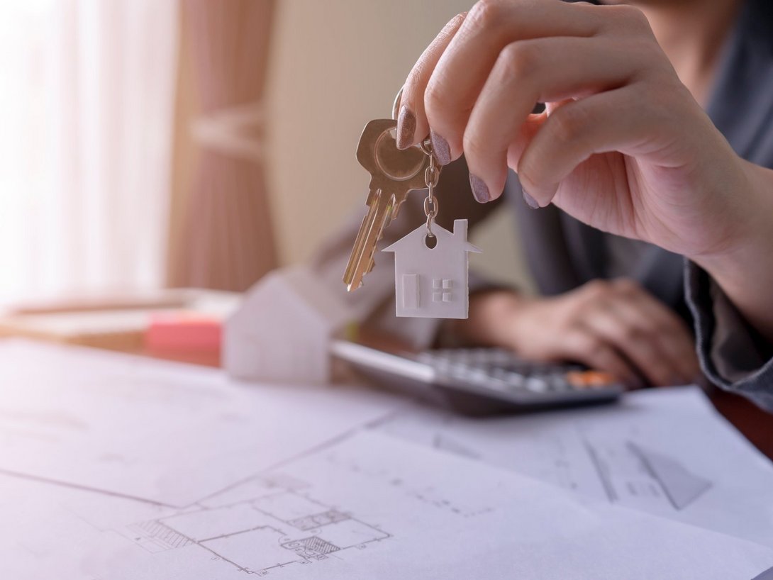 Hand hält Hausschlüssel über Bauplan und Taschenrechner – Symbolbild für Hausbau, Planung und Eigenheim.