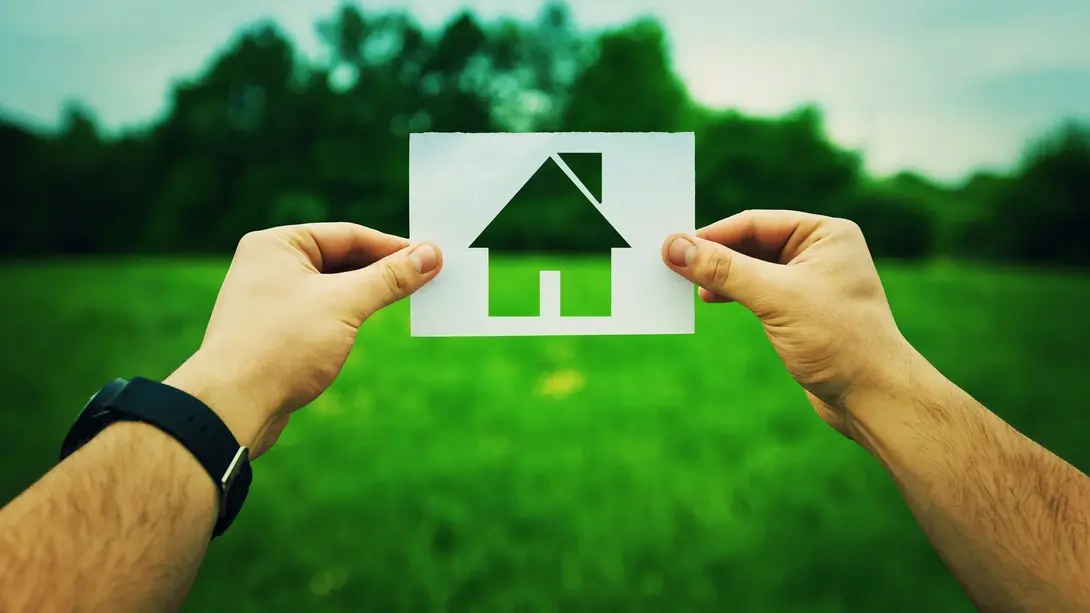 Hände halten ein Haus-Symbol vor einer grünen Wiese als Sinnbild für den Traum vom eigenen Zuhause