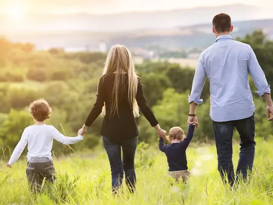 Ein Eigenheim schafft Raum für gemeinsame Zukunft und Lebensqualität. Familie mit zwei Kindern läuft Hand in Hand über eine Wiese – Symbolbild für Familienleben im eigenen Haus.