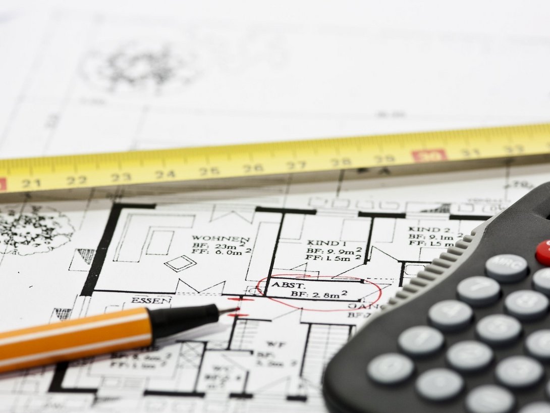 Bauplan mit Maßband, Bleistift und Taschenrechner als Symbol für Planung und Kalkulation einer Neubauimmobilie.