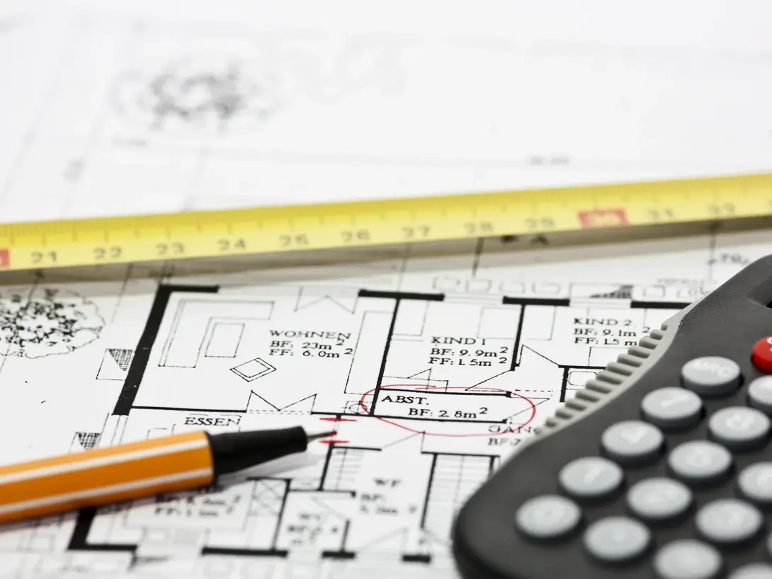 Bauplan mit Maßband, Bleistift und Taschenrechner als Symbol für Planung und Kalkulation einer Neubauimmobilie.