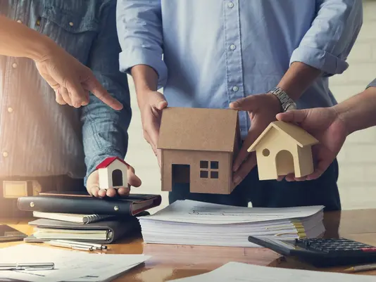 Mehrere Personen planen ein Immobilieninvestment mit Hausmodellen, Bauhelm und Unterlagen auf einem Tisch.