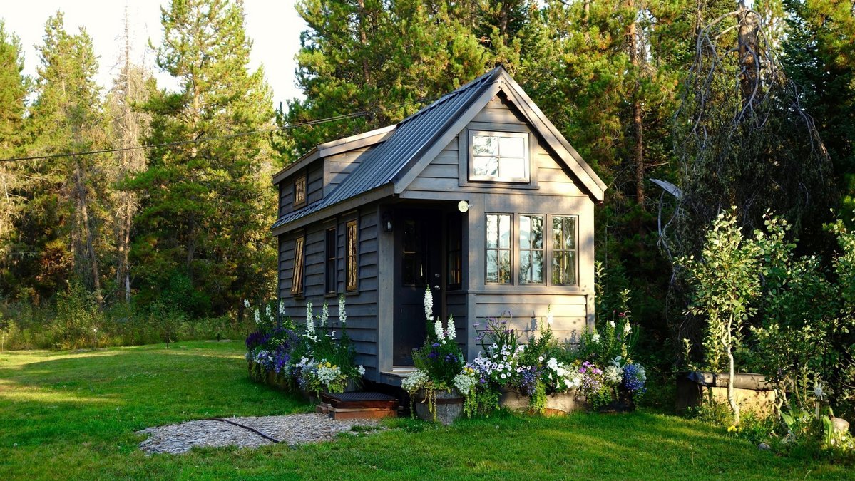 Tiny House aus Holz mit Satteldach, Blumenbeeten und grünem Grundstück am Waldrand