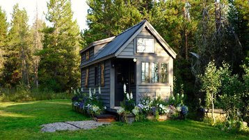 Tiny House aus Holz mit Satteldach, Blumenbeeten und grünem Grundstück am Waldrand