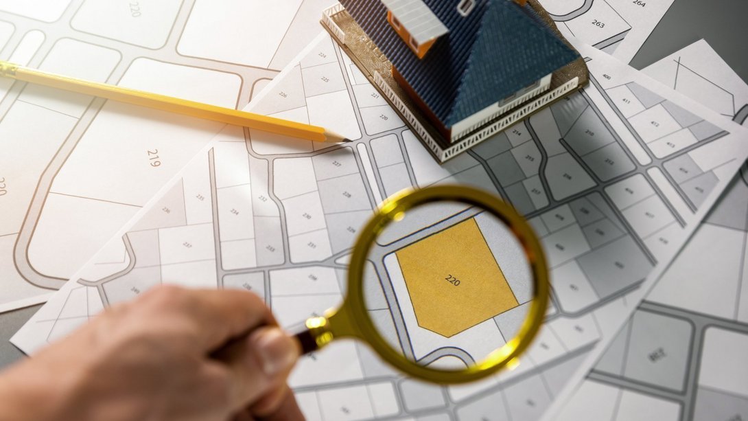 Hand mit Lupe über einem Bebauungsplan, markiertes Grundstück, daneben Modellhaus und Bleistift.