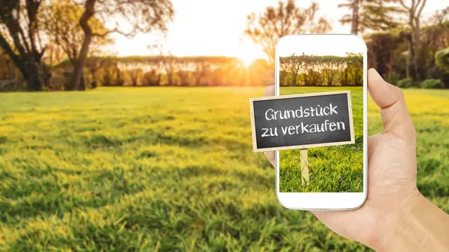 Hand hält ein Smartphone mit Verkaufsschild für ein Grundstück vor einer grünen Wiese bei Sonnenuntergang
