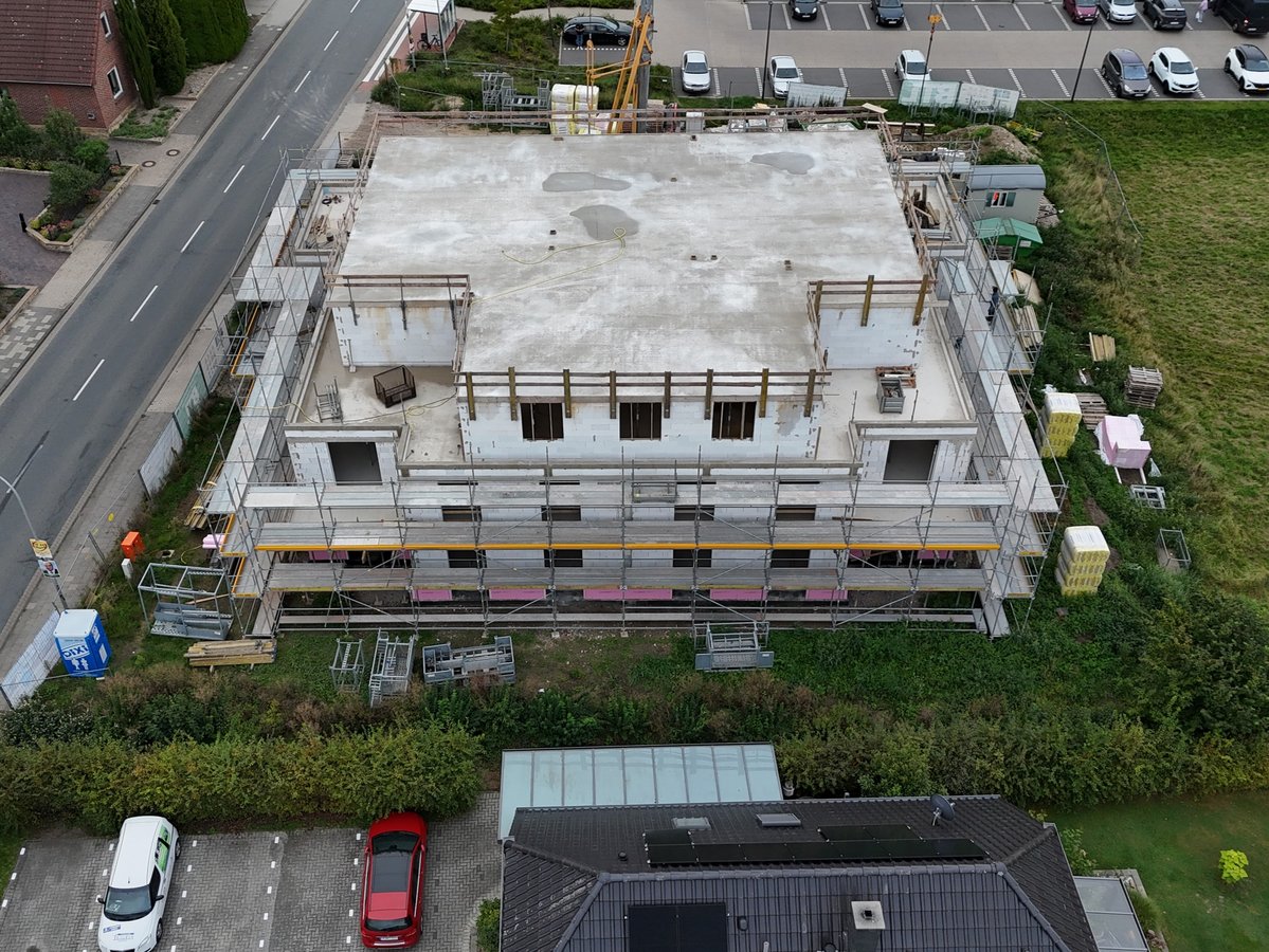 Luftaufnahme eines mehrgeschossigen Rohbaus mit Flachdach und Baugerüst neben einer Straße.