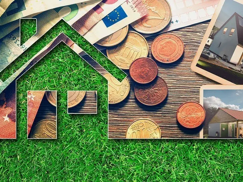 Haus-Symbol aus Gras mit Münzen, Bauunterlagen und Hausbildern als Symbol für Baukosten und Finanzierung