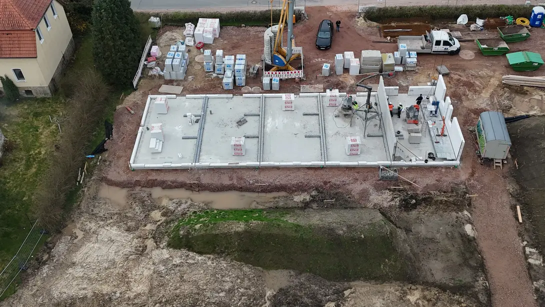 Luftaufnahme einer Hausbaustelle mit Bodenplatte, Baumaterialien, Kran und beginnenden Mauerarbeiten