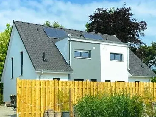 Energieeffizientes Massivhaus in Rhein Modernes Einfamilienhaus mit grauem Satteldach, Photovoltaikanlage, weiß-grauer Fassade und Holzsichtschutzzaun im Vordergrund.