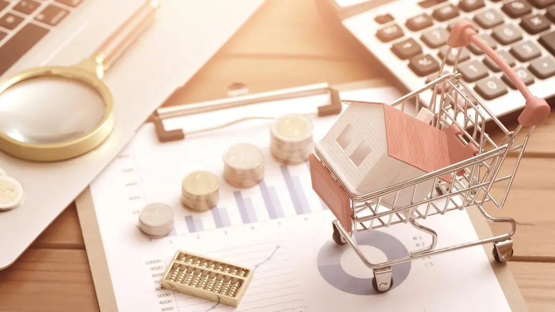Hauskauf und Baufinanzierung planen Miniaturhaus in einem Einkaufswagen neben Münzen, Diagrammen, Taschenrechner und Laptop als Symbol für Immobilienkauf und Baufinanzierung