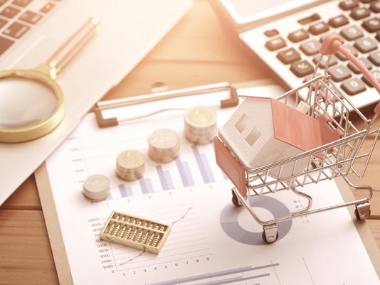 Hausmodell im Einkaufswagen mit Taschenrechner und Finanzunterlagen – Baukosten und Finanzierung beim Hausbau in Westerkappeln planen.
