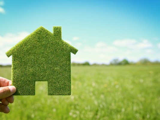 Hand hält ein hausförmiges Symbol aus Gras vor einer grünen Wiese – Sinnbild für nachhaltiges Bauen in Rheine.