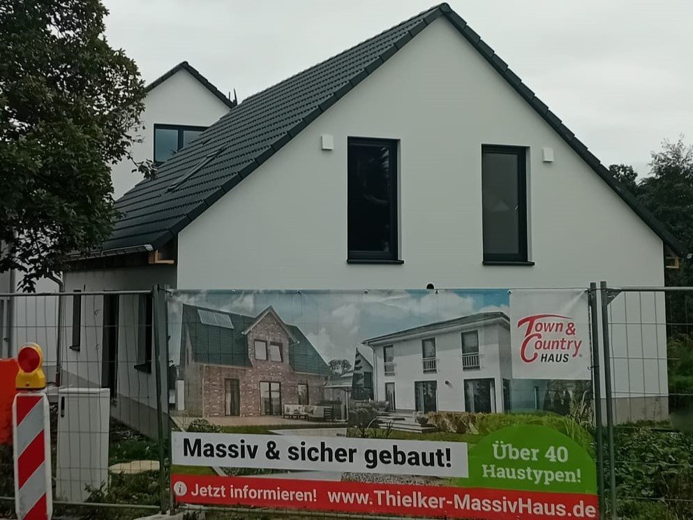Neubau-Einfamilienhaus mit grauem Satteldach hinter einem Bauzaun mit Thielker Massivhaus/Town & Country Haus Werbebanner.